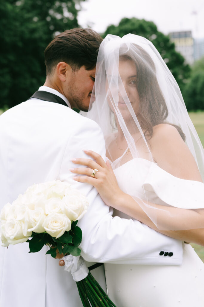 Groom smiles at bride while bride gazes outwards underneath blusher veil.