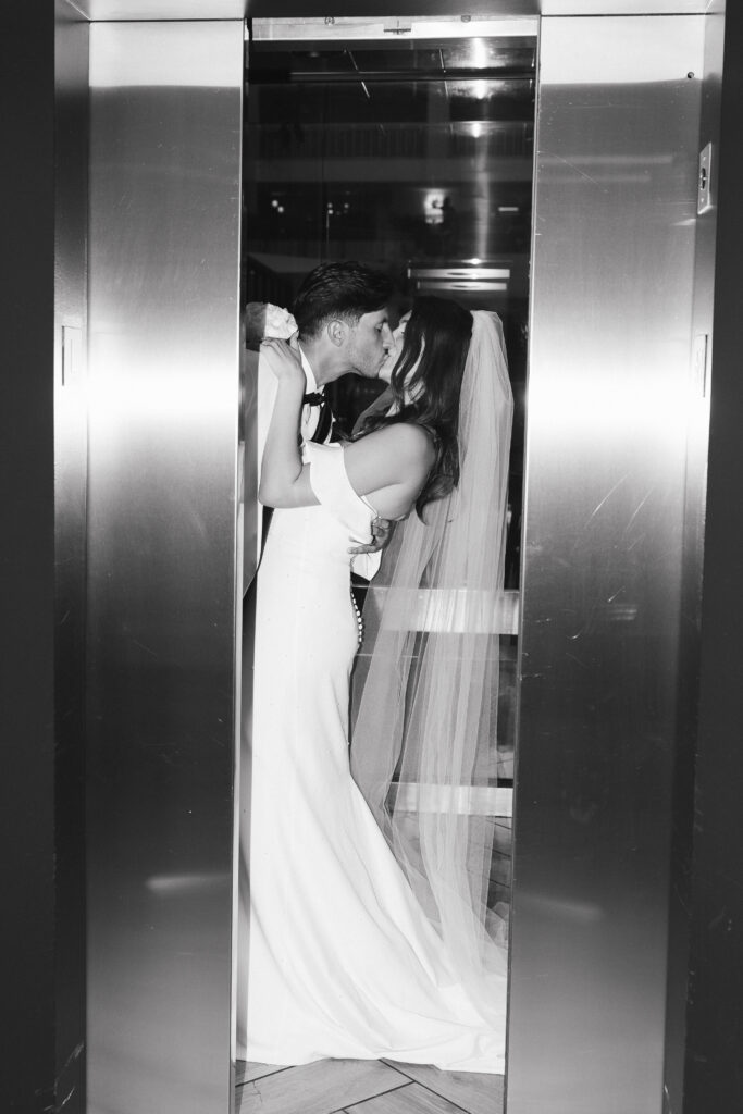 In black and white of bride and groom kissing in an elevator while the doors close.