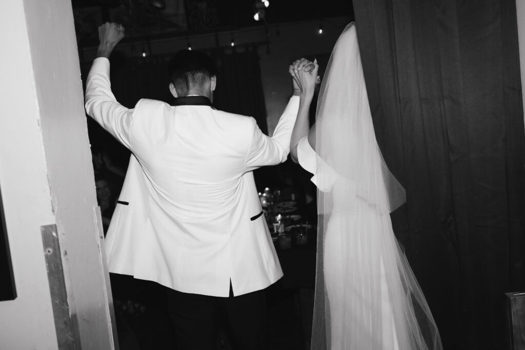 In black and white of bride and groom cheering with hands clasped entering reception space.