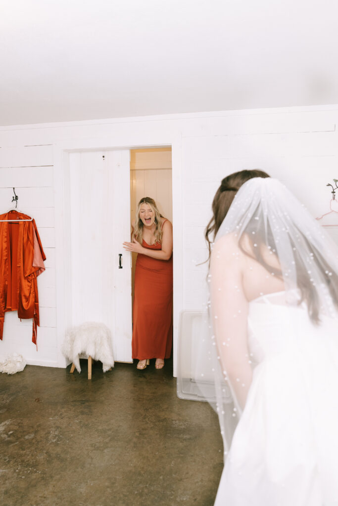 Bridesmaid in an orange dress entering room and seeing bride for the first time.