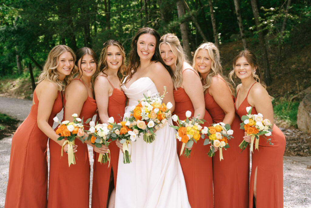 Bride with bridesmaids in orange dresses with white, yellow, and orange flowers.