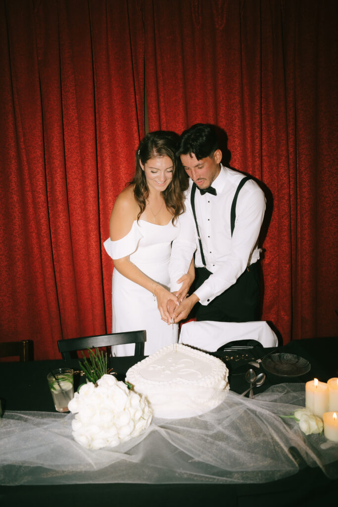 Bride and groom cutting their wedding cake together in front of a red curtain.