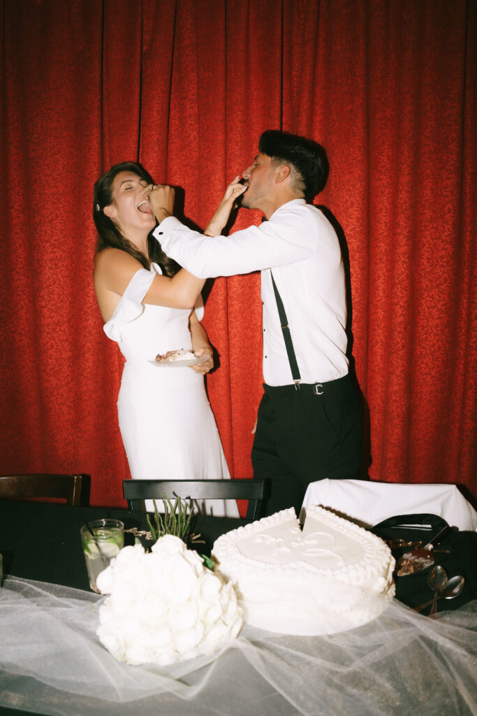 Bride and groom putting cake on each other's faces and laughing in front of a red curtain.