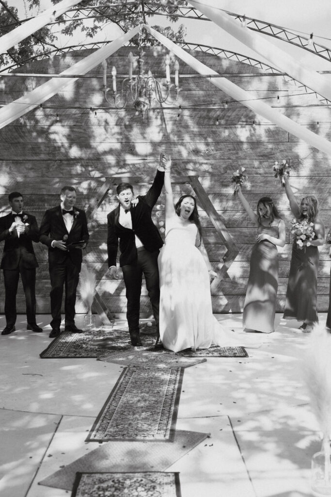 In black and white of bride and groom cheering, clasping raised hands, walking back down the aisle.